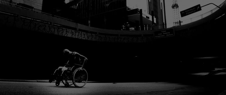 dark image of a man with no legs sat in a wheelchair in a downtown city