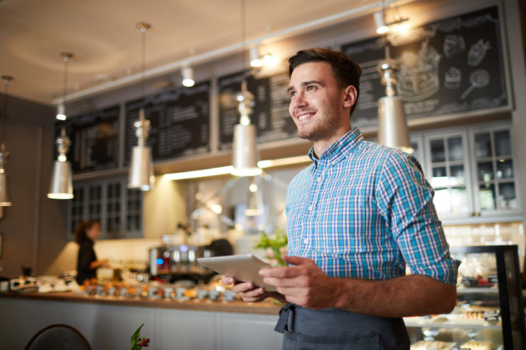 Smiling man holding a tablet in a cafe wearing a blue checked shirt and an apron