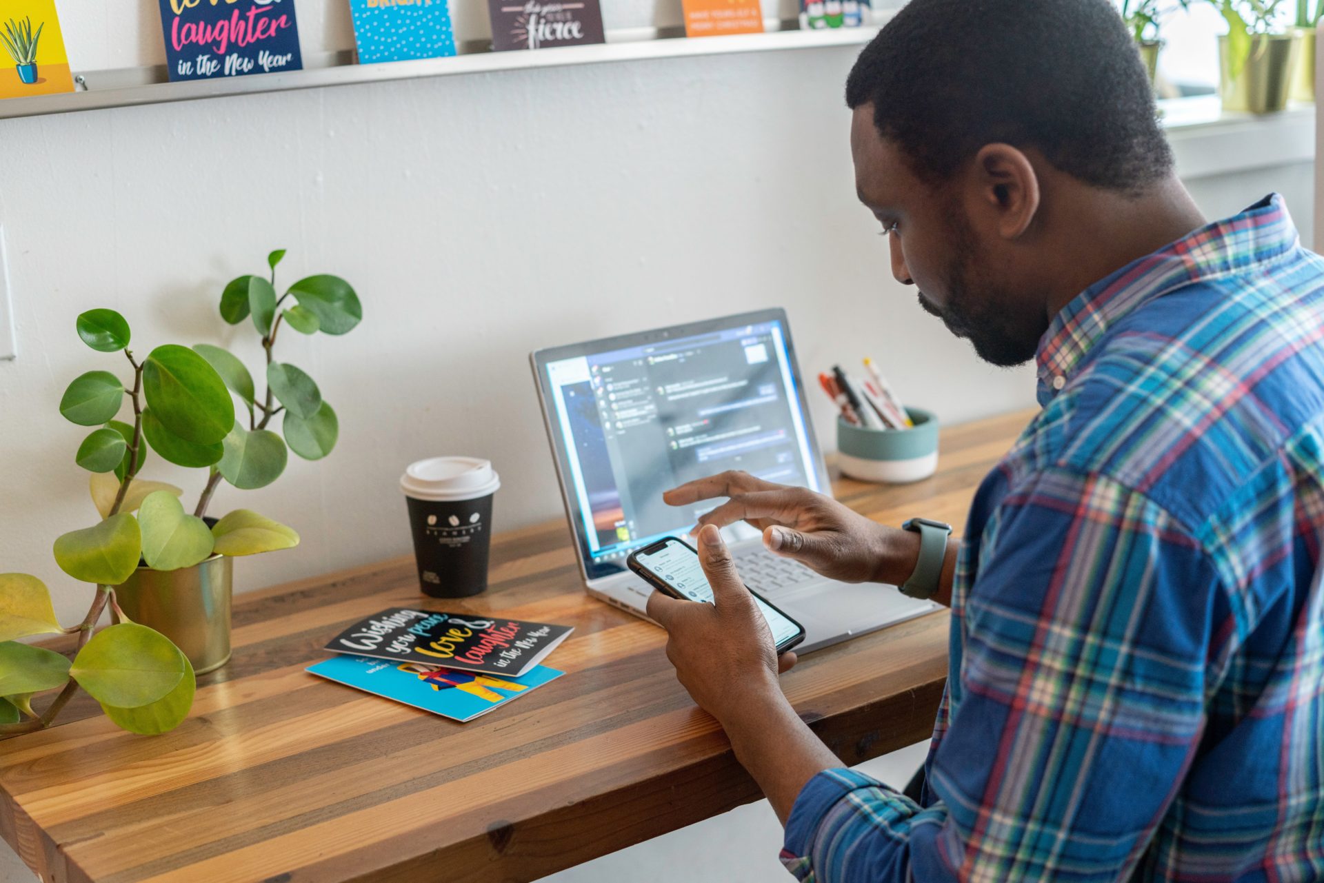 Man hybrid working at desk Man using mobile phone to work and his laptop on a desk with a coffee and cards wearing a multi coloured checked shirt