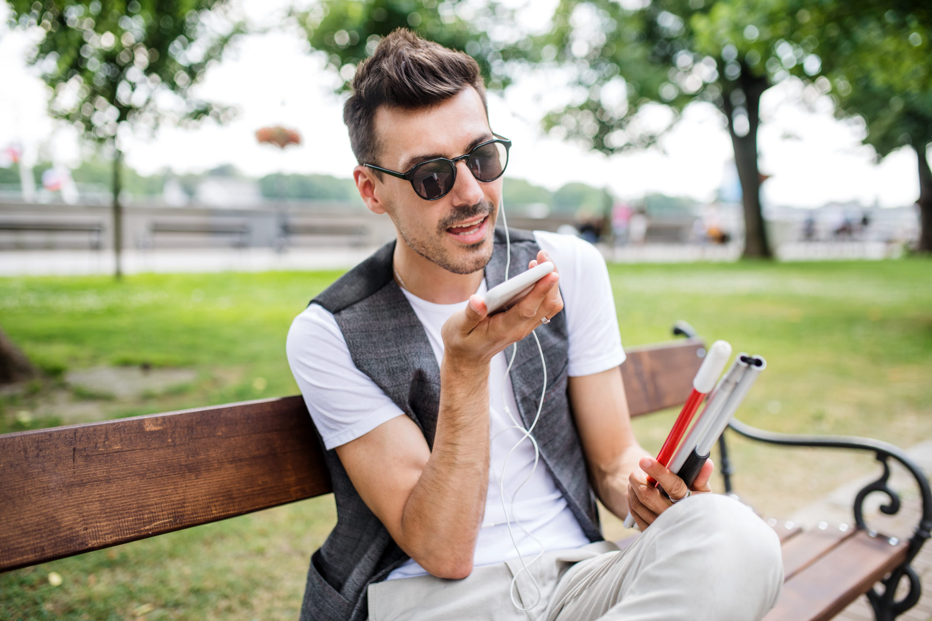 Man talking on mobile phone with earphones wearing sunglasses and a grey waistcoat on a bench outside