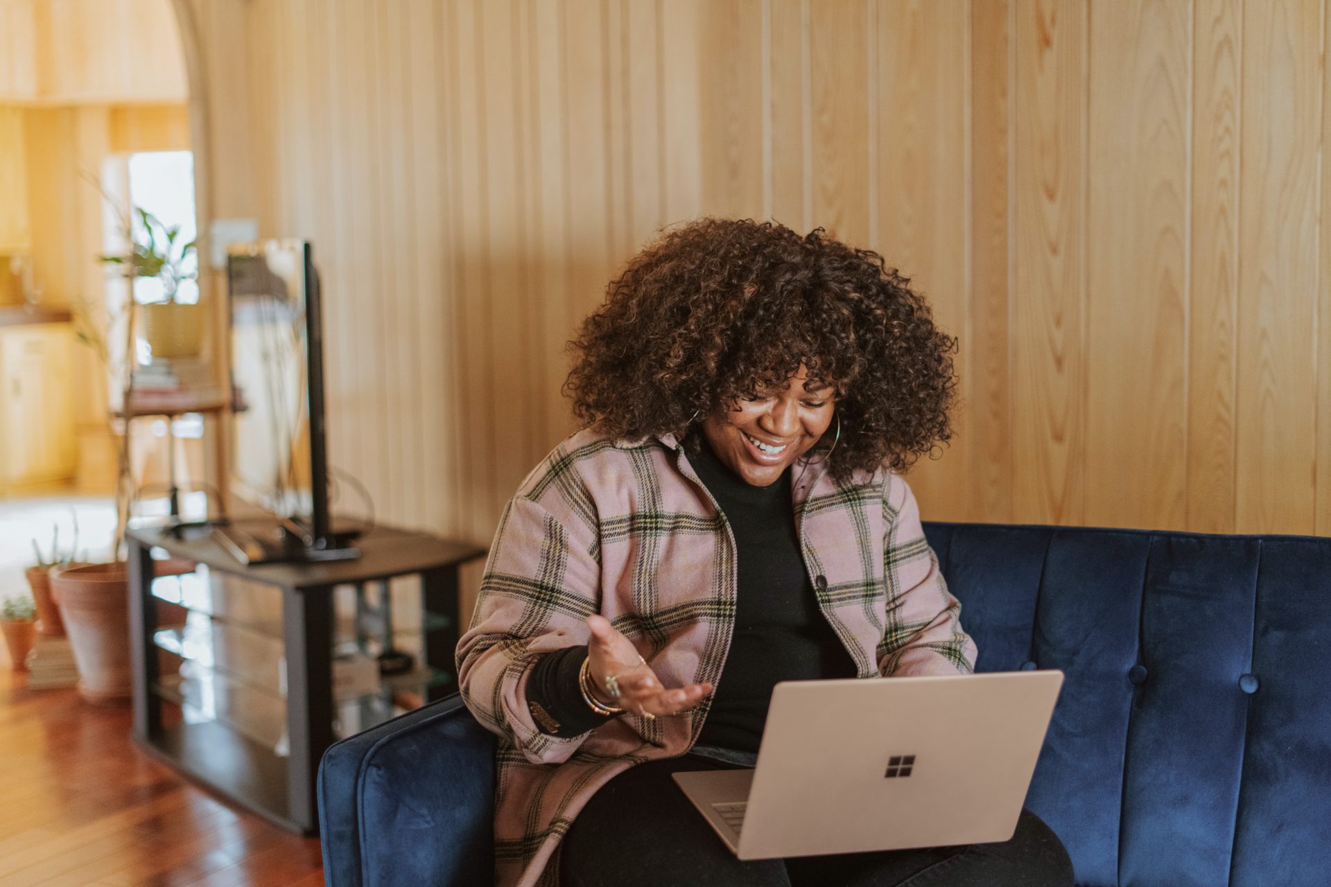 Woman talking on laptop and smiling on blue sofa Woman smiling and working on laptop sat on a blue sofa wearing a pink checked shirt