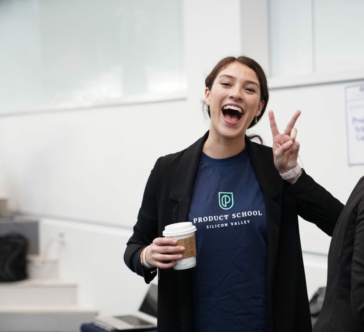 woman looking extremely happy holding a coffee and holding up a peace sign with her hand in an office space