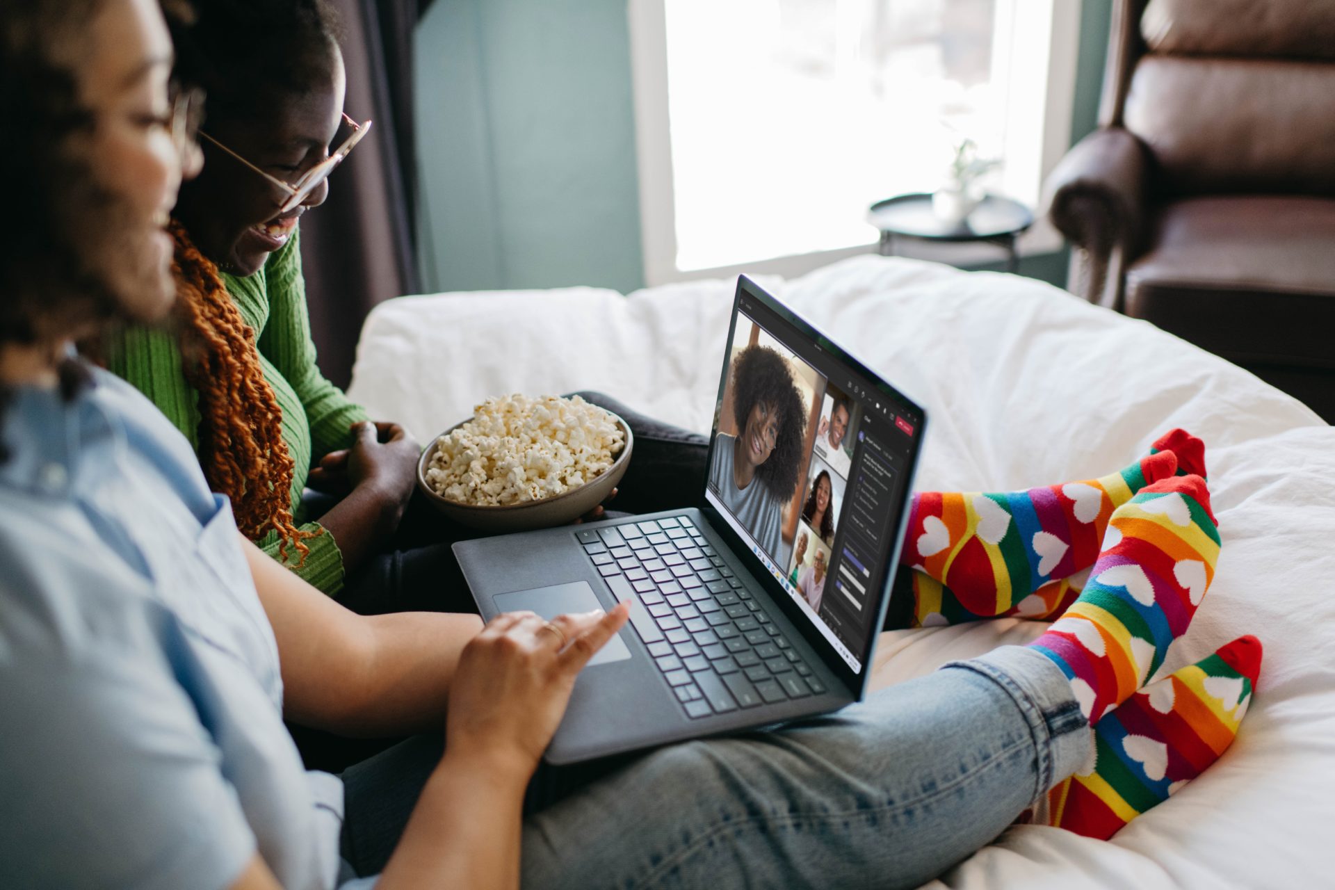 Two woman sat on a bed with popcorn and colourful socks during a Microsoft Teams video call