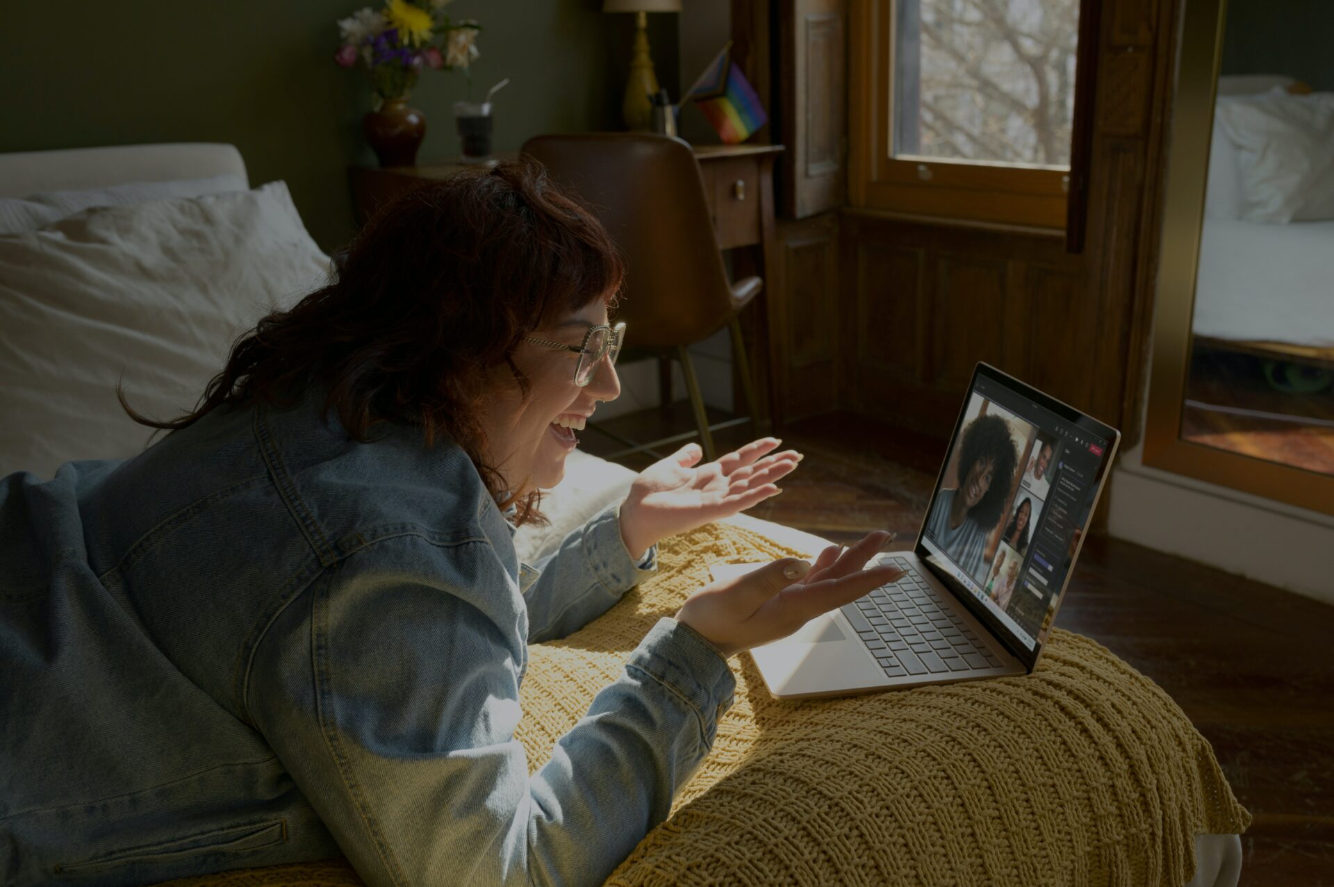 a image of a woman lying on her front on a bed, laughing and smiling at her laptop. on the laptop screen we can see a teams meeting in progress.