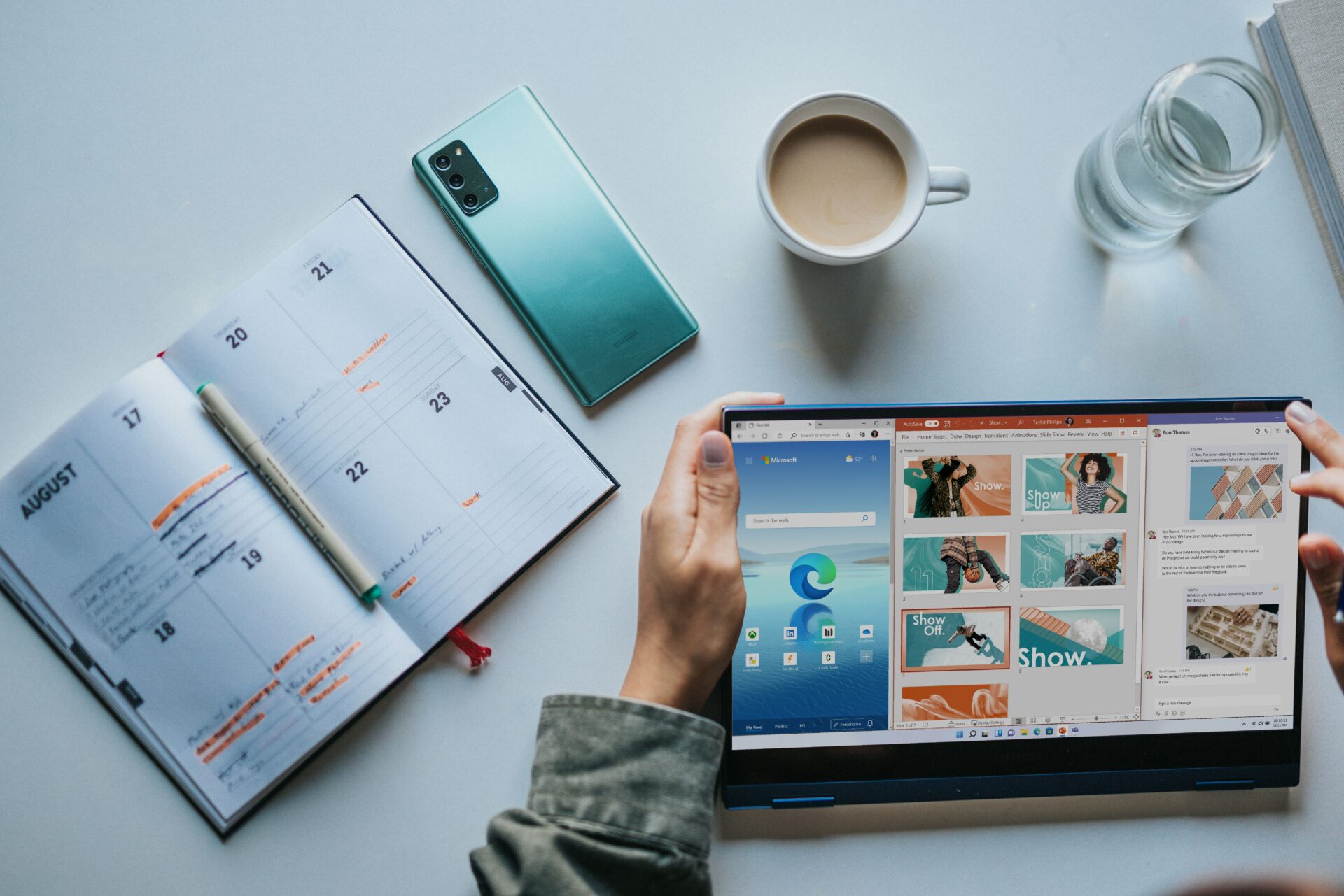 someone holding a tablet with three open windwos - edge, powerpoint and teams. there is an open diary on the table, a mug of tea, a phone face-down and a glass of water