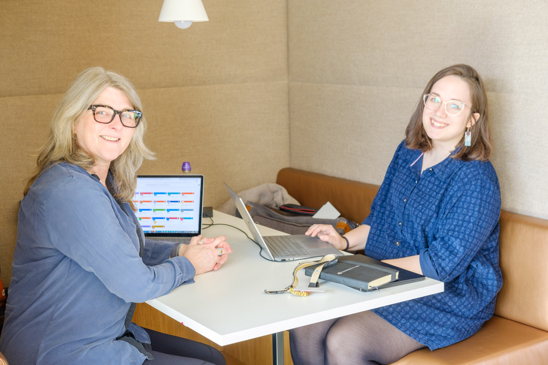 an imge of a woman in a blue top and a person in a blue dress sitting in a both, on either side of a desk. they have laptops open on the table and are smiling at the camera